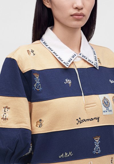 Femme portant une chemise beige et bleu marine rayée avec des armoiries brodées, des mots, et un col blanc avec une bordure à motif bleu.