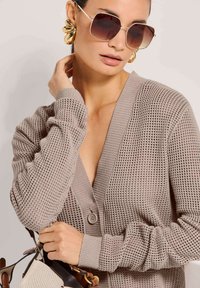 Beige gestrickte Strickjacke mit V-Ausschnitt und Knopfverschluss, die ein strukturiertes Muster aufweist. Übergroße Sonnenbrillen und goldene Creolen runden den Look ab.