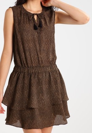 Vestido marrón sin mangas con patrón geométrico, cintura elástica, falda con volantes y lazo con borlas en el escote, usado por una mujer.