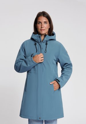Vrouw draagt een knielange blauwe winterjas met een kap, één hand past de voorste rits aan, de andere in een zijzak.