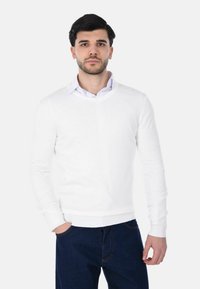 Giovane uomo con capelli scuri che indossa un maglione bianco a V sopra una camicia con colletto e jeans di denim scuri, in piedi contro uno sfondo bianco semplice.
