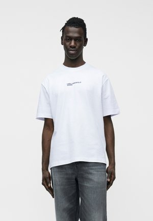 Joven con camiseta blanca de manga corta con el texto "KARL LAGERFELD JEANS" y vaqueros grises desgastados, de pie contra un fondo liso.