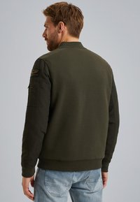 Veste bomber olive avec un corps texturé, des manches lisses, des poignets côtelés et une poche latérale ; présente un patch sur le bras gauche.
