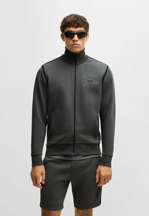 Giacca sportiva grigio scuro con colletto alto, chiusura a zip e tasche frontali. Abbinata a pantaloni corti coordinati, entrambi in tessuto liscio.