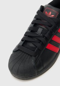 Czarny skórzany sneaker z trzema czerwonymi paskami, okrągły nosek z teksturowaną gumową osłoną i czarnymi sznurowadłami. Prosty, klasyczny sportowy design.