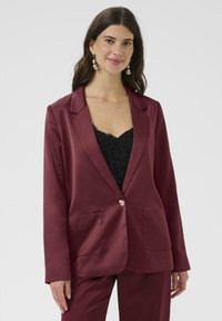 Donna che indossa un blazer satinado bordeaux e pantaloni abbinati su un top in pizzo nero, in piedi con un leggero sorriso e orecchini d'argento.