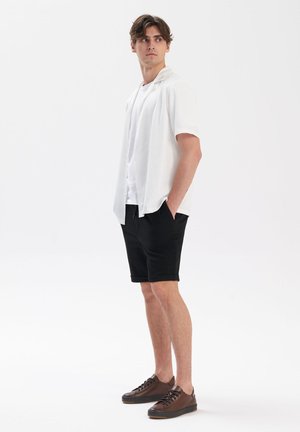 Biała koszula z krótkim rękawem na białej koszulce, w zestawie z czarnymi szortami i brązowymi sneakersami. Koszula ma luźny krój i otwarty kołnierz.