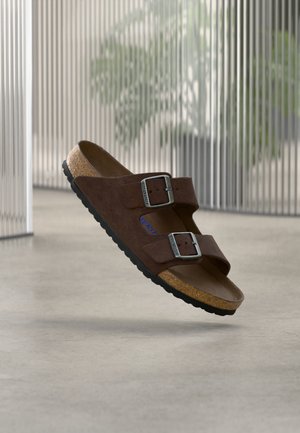 Barna velúr szandál két állítható fém csattal és parafa talpbetéttel, sima padló felett függ.