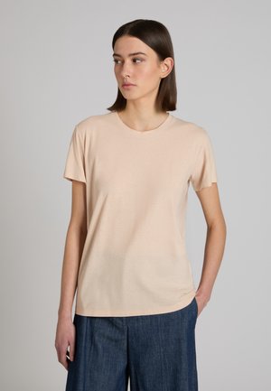 Młoda kobieta o prostych brązowych włosach, ubrana w beżowy T-shirt z krótkim rękawem i ciemnoniebieskie spodnie, stojąca z jedną ręką w kieszeni.