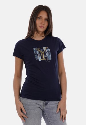 Donna con capelli castani lunghi che indossa una maglietta blu scuro con una grande "Y" gialla stilizzata e un design astratto blu, abbinata a jeans grigio chiaro.