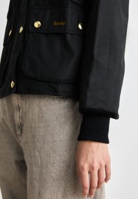 Zwarte gewaxte jas met gouden details, voorzien van dubbele voorzakken, geribbelde zwarte manchetten en de merknaam "Barbour" geborduurd op de borstzak.