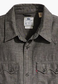 Camicia western in denim grigio con colletto a punta, bottoni a pressione, due tasche sul petto e design a vestibilità rilassata. Etichetta del marchio visibile all'interno del colletto.