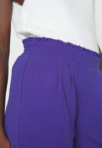 Purple high-waisted trousers with a ruffled waistband and front pleats. Made of smooth fabric, featuring a fitted silhouette.