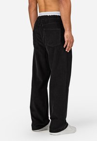 Czarne spodnie z korduroy o luźnym kroju, z elastycznym paskiem i dwiema tylnymi kieszeniami. Faktura materiału jest prążkowana. Nosi się je z białymi butami.