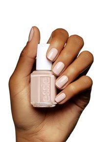 Bouteille de vernis à ongles avec un capuchon blanc et un vernis de couleur rose beige tenue dans une main avec des ongles manucurés assortis à la couleur du vernis. Texture lisse.