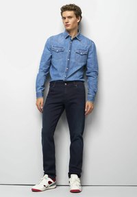 Hellblaues Jeanshemd mit zwei Brusttaschen, dunkelmarineblauen Hosen, weißen Sneakers mit roten Akzenten. Modell steht vor einem neutralen Hintergrund.
