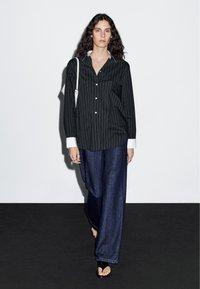 Black and white striped button-up shirt with long sleeves and wide cuffs, paired with loose blue jeans and open-toed sandals.