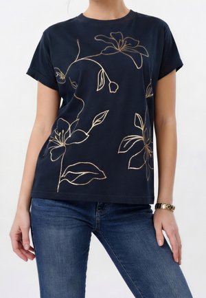Kvinde iført en sort kortærmet skjorte med et guld blomsterline mønster og blå jeans, stående med hænderne nær hofterne.