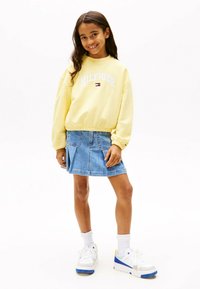 Sudadera amarilla clara con el logo "HILFIGER", combinada con una falda de mezclilla azul. Zapatillas blancas con detalles en azul y amarillo.