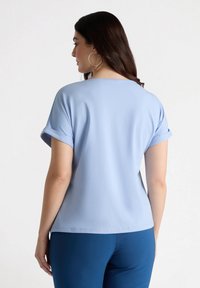 Jasnoniebieska bluzka z krótkim rękawem z podwiniętymi mankietami, okrągłym dekoltem i gładką fakturą. Połączona z ciemnoniebieskimi spodniami. Prosty design.