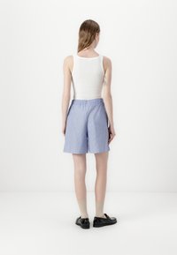 Kobieta stojąca tyłem, ubrana w białą, bezrękawnikową bluzkę, luźne niebieskie szorty w paski, beżowe skarpetki i czarne buty na białym tle.