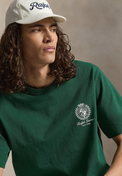 Junger Mann mit langen lockigen Haaren, der eine weiße Ralph-Cap und ein dunkelgrünes Ralph Lauren T-Shirt mit Logo auf der linken Brust trägt.