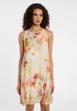 Vrouw met krullend donker haar, gekleed in een mouwloze beige jurk met roze, oranje en witte bloemenpatronen, staande tegen een effen witte achtergrond.