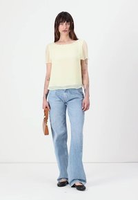Chemisier léger jaune clair avec manches courtes, design superposé, associé à un jean évasé bleu clair et des chaussures plates noires. Tenue d'un petit sac à main marron.