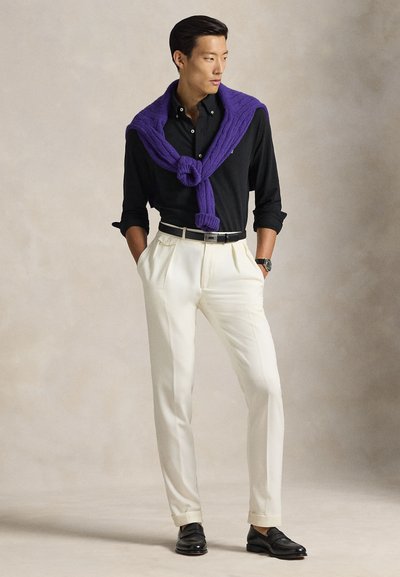 Vyras, dėvintis juodą marškinių ir violetinį megztinį per pečius, baltas sijonas su raukiniais, juodas diržas ir juodi batai, stovintis su rankomis kišenėse.