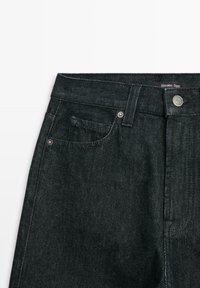 Tmavé džíny mají vysoký pas s knoflíkovým zapínáním, klasické přední kapsy a diskrétní prošívání, prezentované na bílém pozadí.