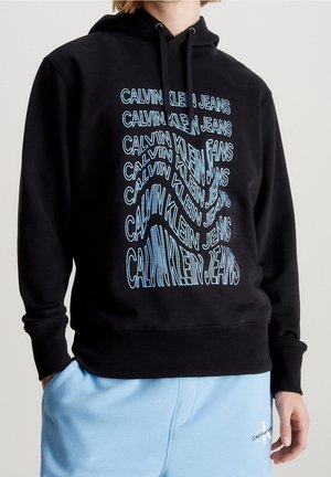 Osoba nosząca czarną bluzę z kapturem z powtarzającym się niebieskim napisem „Calvin Klein Jeans” oraz jasnoniebieskie spodnie dresowe z małym logo na lewej nodze.