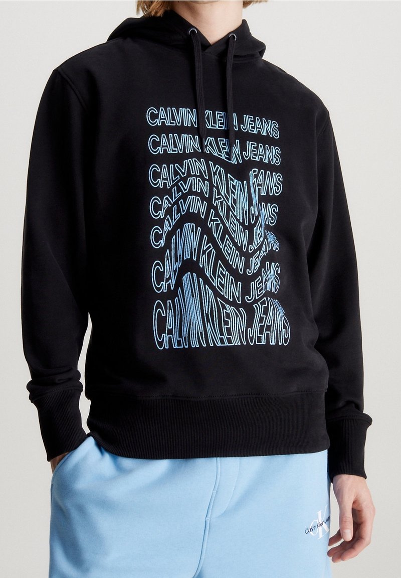 Personne portant un sweat à capuche noir avec le texte bleu répété « Calvin Klein Jeans » et un pantalon de survêtement bleu clair avec un petit logo sur la jambe gauche.