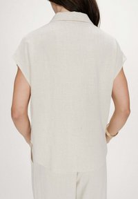 Mujer vistiendo una camisa beige de lino suelta y de manga corta, y pantalones a juego, vista desde atrás con las manos en los bolsillos y pulseras doradas en la muñeca.