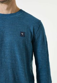 Blauwe gebreide trui met een ronde hals. Heeft een klein stoffen logo op de linkerkant. Zachte textuur en aansluitend ontwerp.