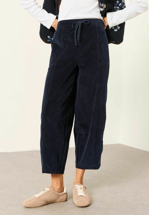 Frau trägt dunkelblaue, weit geschnittene Cordhose mit Kordelzug in der Taille und beige Schnürfreizeitschuhe, steht auf einem grauen Boden.