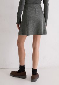 Femme portant une jupe grise, un haut à manches longues assorti, des chaussettes noires et des mocassins marron, debout contre un mur blanc.