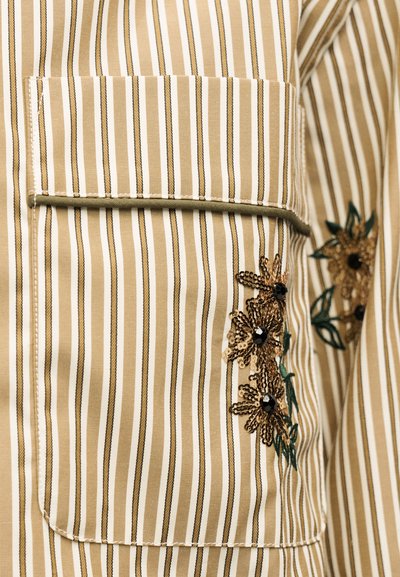 Υφασμα με κάθετες ρίγες σε μπεζ και άσπρο χρώμα με τσέπη, διακοσμημένο με κεντήματα από παγιέτες σε χρυσό και μαύρους χάντρες σε σχέδιο λουλουδιών.