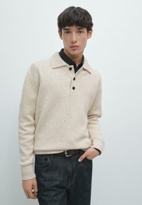 Beżowy sweter dzianinowy z kołnierzem polo, plisą na guziki i ściągaczami przy rękawach, zestawiony z ciemnoniebieskimi dżinsami i brązowym paskiem. Prosty design.