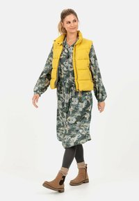 Gul polstret vest over en grøn blomsterkjole. Brune ankelstøvler med strikkede manchetter. Mørke leggings. Enkel baggrund, model går.