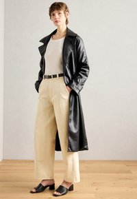 Cappotto in finta pelle nera, pantaloni ampi beige, sandali neri con tacco a blocco. Tessuto morbido, grande colletto, dettaglio con fibbia dorata.