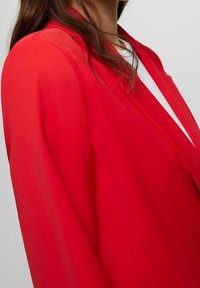 Rød blazer med en glat tekstur, løs pasform og sjalskrave. Stoffet virker let og en smule gennemsigtigt.