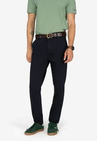 Pantaloni scuri blu navy con una vestibilità slim, realizzati in tessuto misto cotone, dotati di tasche laterali e una cintura in pelle marrone. Le sneakers verdi completano il look.