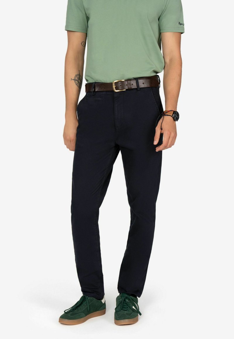 Pantaloni scuri blu navy con una vestibilità slim, realizzati in tessuto misto cotone, dotati di tasche laterali e una cintura in pelle marrone. Le sneakers verdi completano il look.