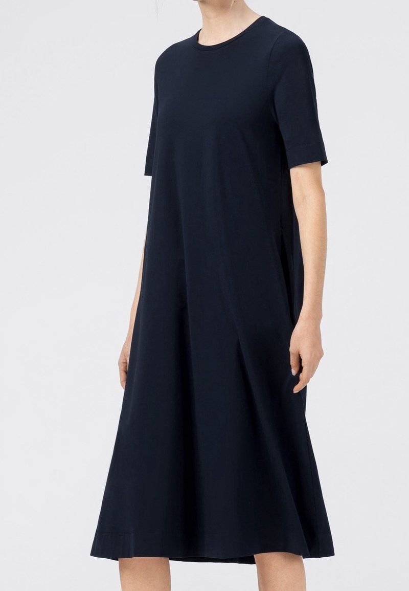 Femme portant une robe noire ample à manches courtes et longueur genou avec un col rond sur un fond uni.