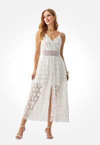 White lace dress featuring a V-neck, adjustable straps, mid-length design, floral pattern, and a front slit. Accented with a crochet waistband.