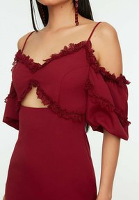 Femme portant une robe bordeaux à fines bretelles, manches bouffantes décolletées, bordure en dentelle et petite découpe à l'avant.