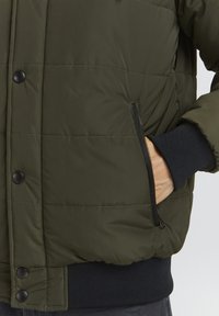 Giacca puffer verde oliva con sezioni trapuntate, bottoni automatici neri e una tasca laterale con zip. Polsini e vita a coste in blu navy.