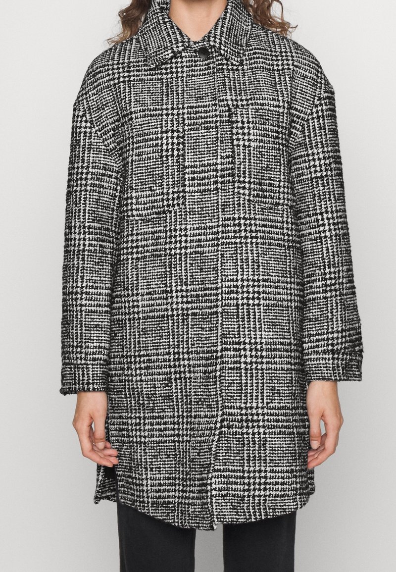 Femme portant un manteau à motif pied-de-poule noir et blanc avec col et boutons sur le devant, debout les bras détendus le long du corps.