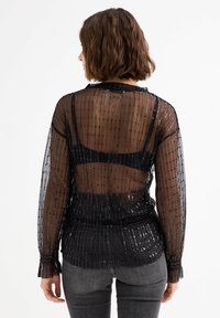 Blusa negra transparente con rayas verticales y sutiles lentejuelas, de manga larga y ajuste holgado. Vista trasera que muestra un diseño en capas.