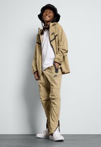 Fato de treino bege da Nike com uma jaqueta com fecho, detalhes em preto contrastantes e calças a condizer com zíperes nos tornozelos. Ténis brancos.